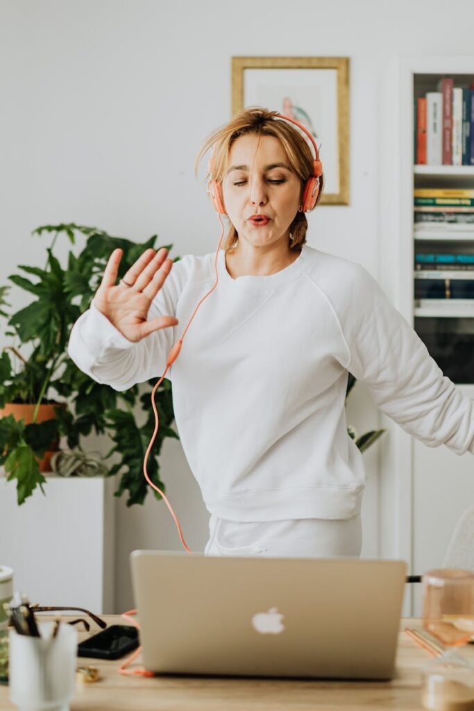 pexels photo 5899248 5899248 An adult woman dancing joyfully in her home office, wearing headphones and using a laptop.