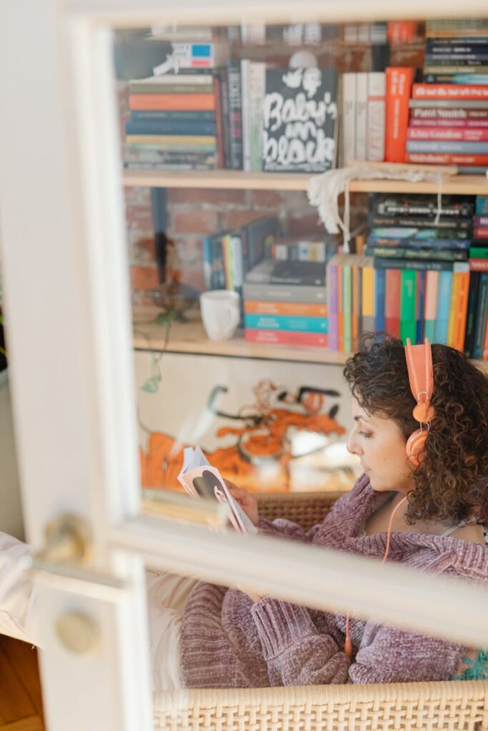 pexels photo 6633272 6633272 Woman relaxing indoors with book and headphones, near a bookshelf.